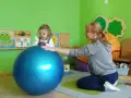 Dziecko w okularach ćwiczy z fizjoterapeutką na dużej, niebieskiej piłce w sali przedszkolnej.