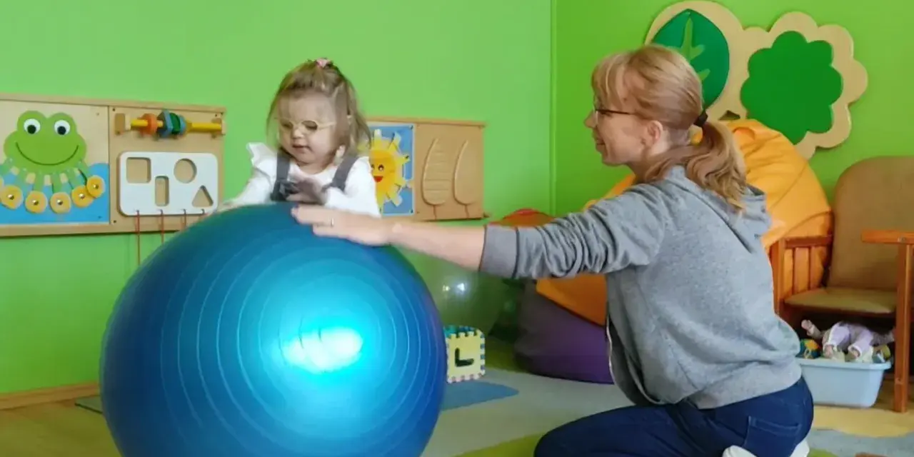 Dziecko w okularach ćwiczy z fizjoterapeutką na dużej, niebieskiej piłce w sali przedszkolnej.