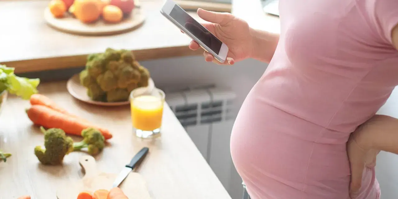 Ciężarna kobieta zmagająca się z brakiem apetytu w ciąży, szuka w telefonie przepisu na zdrowe jedzenie. Na blacie warzywa i sok.