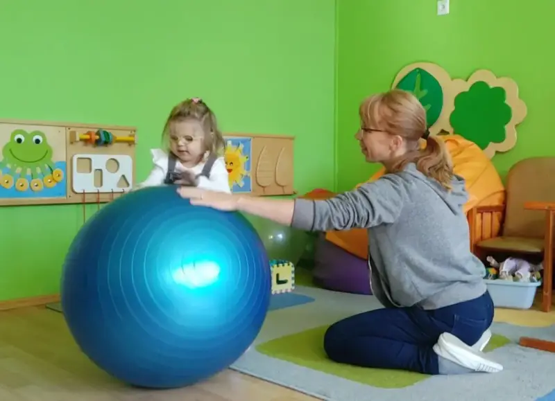 Dziecko w okularach ćwiczy z fizjoterapeutką na dużej, niebieskiej piłce w sali przedszkolnej.
